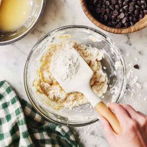 Mixing flour into cookie dough in a glass bowl with chocolate chips nearby