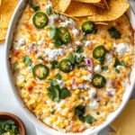 Golden bubbly Mexican street corn dip in white baking dish topped with melted cheese, fresh cilantro, sliced jalapeños, red onions, and crumbled feta with tortilla chips on the side
