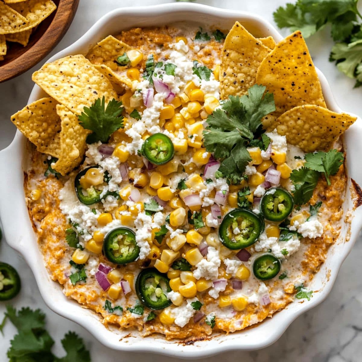 Close-up view of creamy corn dip in white serving bowl garnished with yellow corn kernels, fresh cilantro, jalapeño slices, purple onions, and white feta cheese with golden tortilla chips arranged around the bowl