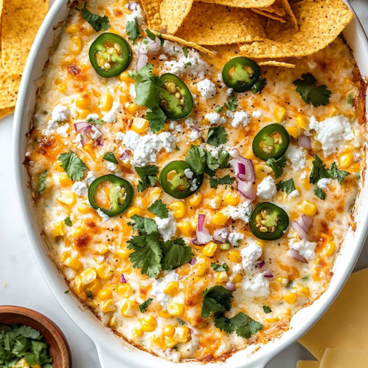 Golden bubbly Mexican street corn dip in white baking dish topped with melted cheese, fresh cilantro, sliced jalapeños, red onions, and crumbled feta with tortilla chips on the side