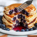 Blueberry pancakes stack being cut with fork showing fluffy interior with bursted blueberries and syrup on white plate with fresh berries