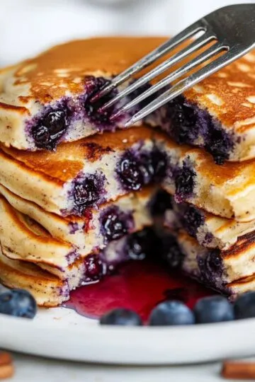 Blueberry pancakes stack being cut with fork showing fluffy interior with bursted blueberries and syrup on white plate with fresh berries