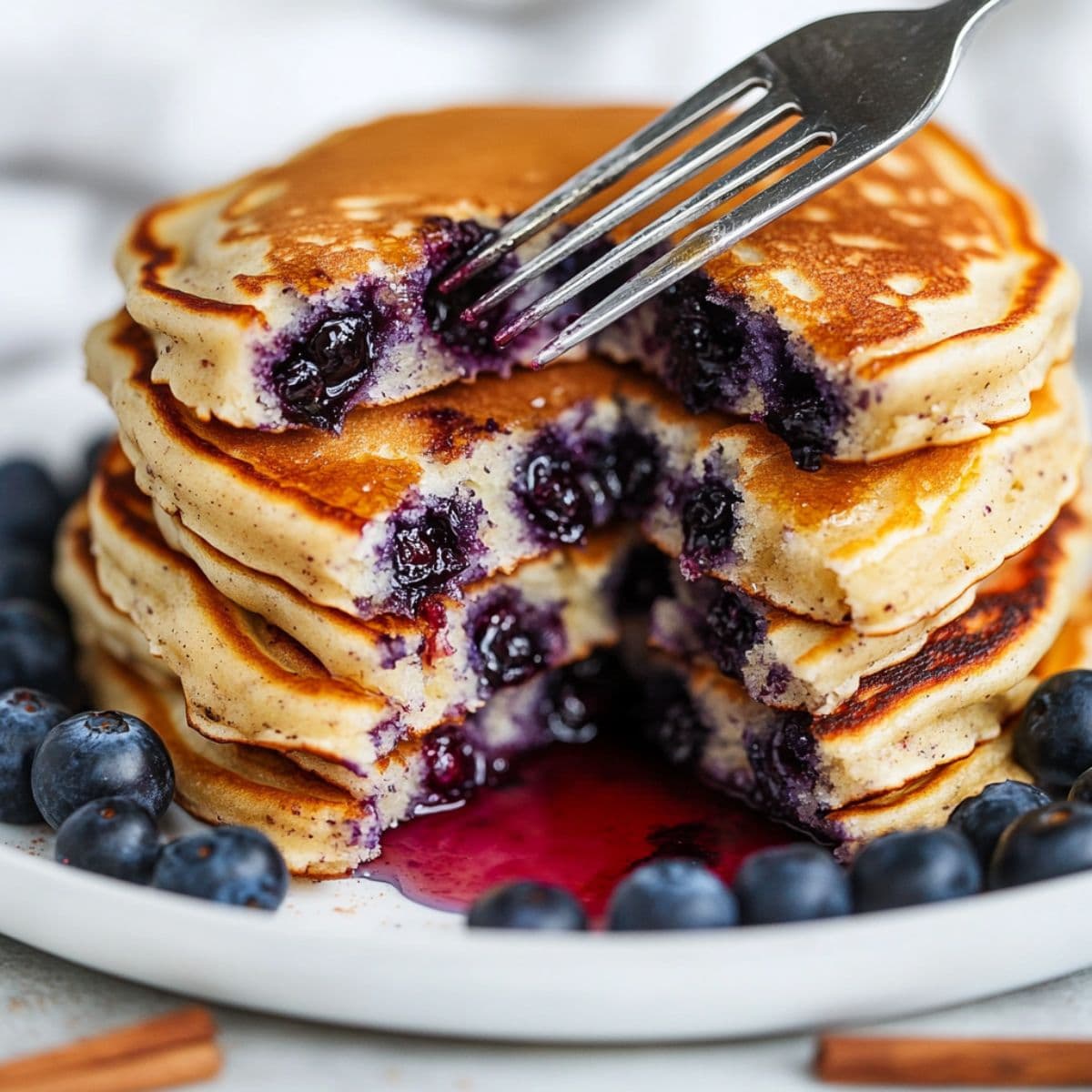 Blueberry pancakes stack being cut with fork showing fluffy interior with bursted blueberries and syrup on white plate with fresh berries