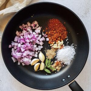 Garlic broccoli stir fry aromatics cooking in dark pan with diced red onions, minced garlic cloves, fresh ginger and colorful spices for aromatic base