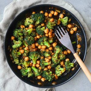 Garlic broccoli stir fry with chickpeas finished dish in dark pan showing vibrant green broccoli florets, golden chickpeas and glossy sauce with wooden spoon