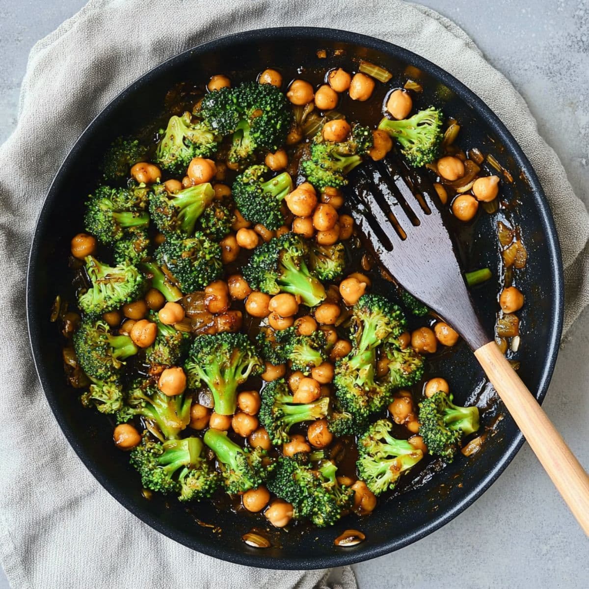 Garlic broccoli stir fry with chickpeas finished dish in dark pan showing vibrant green broccoli florets, golden chickpeas and glossy sauce with wooden spoon