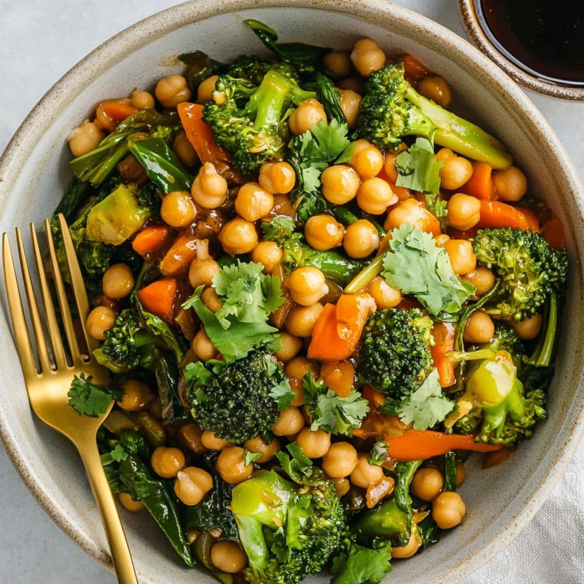 Garlic broccoli stir fry with chickpeas served in ceramic bowl with gold fork, showing vibrant vegetables, chickpeas and fresh cilantro garnish for healthy meal presentation