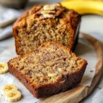 Healthy banana bread close-up showing incredibly moist texture with visible banana pieces and golden brown crumb on wooden board with fresh bananas