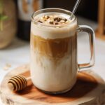 Perfect honey oat milk latte in clear glass mug showing beautiful golden layers, topped with honey drizzle and foam, styled with honey dipper on wooden board