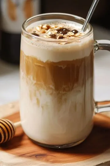 Perfect honey oat milk latte in clear glass mug showing beautiful golden layers, topped with honey drizzle and foam, styled with honey dipper on wooden board
