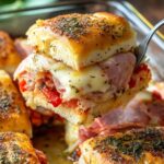 Close-up of hot Italian slider sandwich with melty cheese, ham, roasted red peppers, and herbs, being lifted from a baking dish – golden and appetizing.