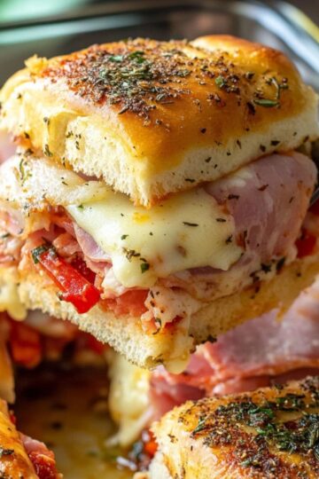 Close-up of hot Italian slider sandwich with melty cheese, ham, roasted red peppers, and herbs, being lifted from a baking dish – golden and appetizing.
