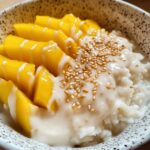 Mango sticky rice recipe showing coconut sticky rice with fresh yellow mango slices, coconut sauce drizzle and sesame seeds in speckled bowl