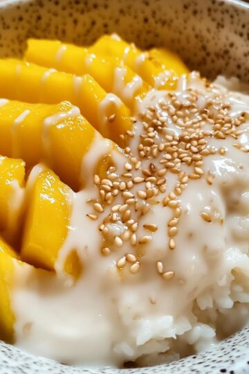 Mango sticky rice recipe showing coconut sticky rice with fresh yellow mango slices, coconut sauce drizzle and sesame seeds in speckled bowl