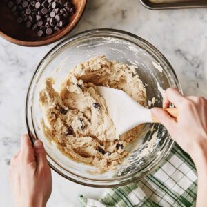 Hands using white spatula to mix thick chocolate chip cookie dough in glass bowl, with chocolate chips scattered throughout and wooden bowl of chocolate chips visible in background