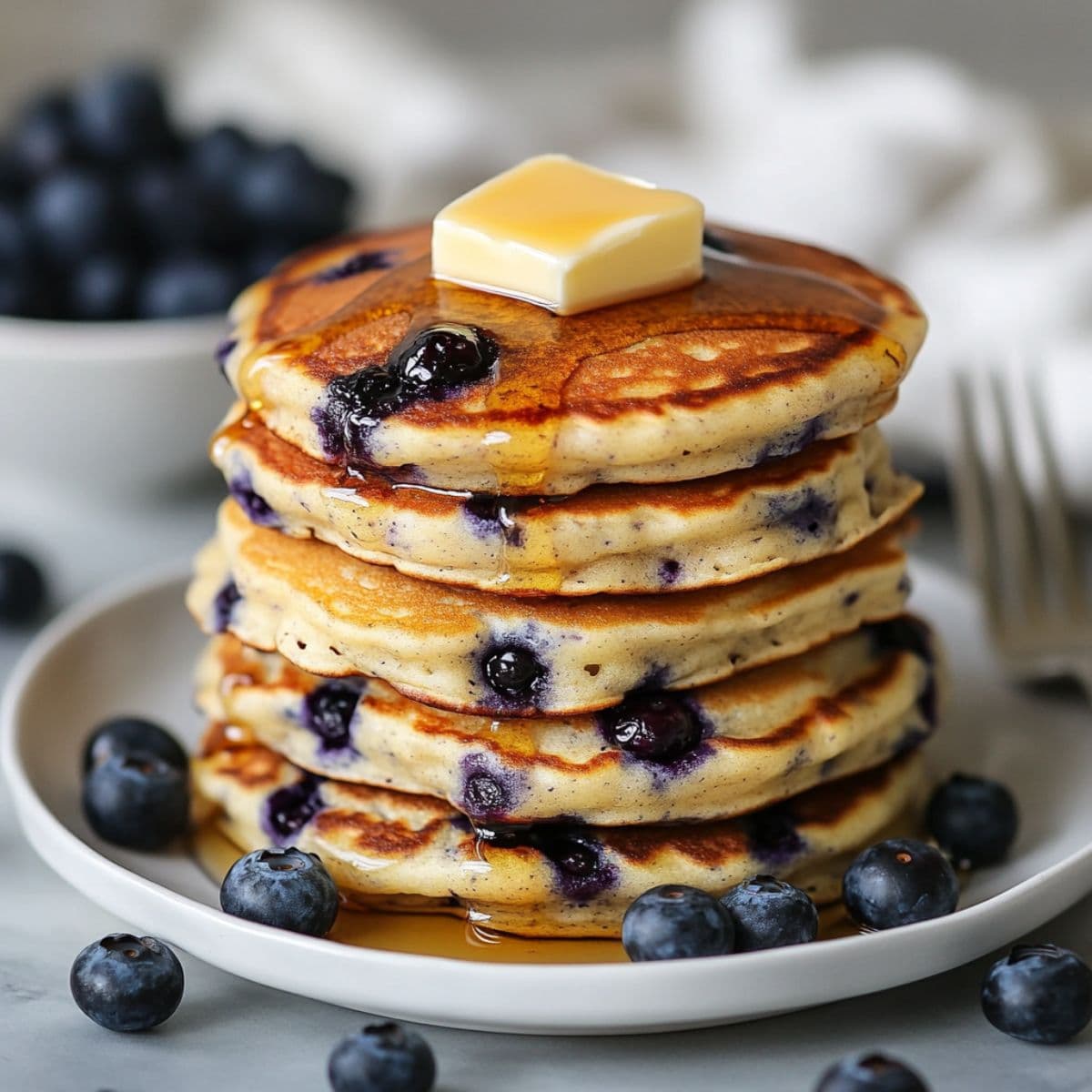 Blueberry pancakes perfect stack with butter on top and maple syrup drizzle surrounded by fresh blueberries on white plate