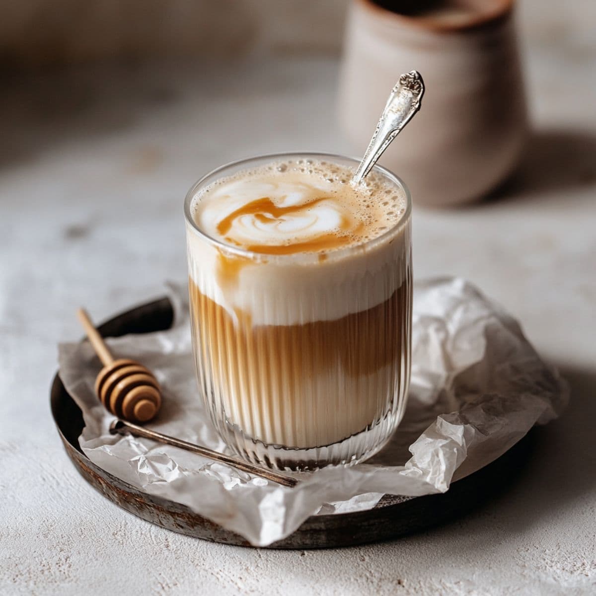 Perfect honey oat milk latte with golden honey swirl on foam, served in elegant ribbed glass with vintage spoon and honey dipper on ceramic plate, ready for serving