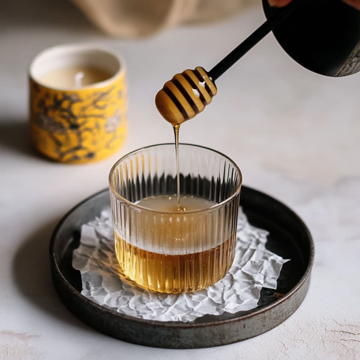 Honey being drizzled from wooden honey dipper into elegant glass cup with layered honey oat milk latte, styled on dark ceramic plate showing the first step of latte preparation