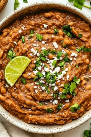 Refried beans recipe showing creamy homemade Mexican beans garnished with fresh cilantro, lime wedge and cotija cheese in rustic bowl