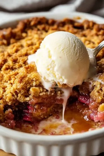 Close-up of a rhubarb crisp topped with melting vanilla ice cream in a white baking dish.