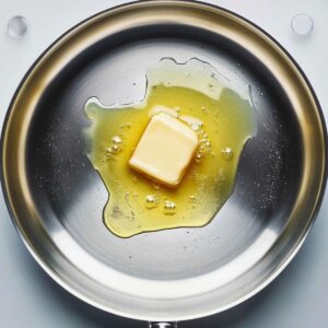 Top-down view of a cube of butter melting in a stainless steel pan on a white background.