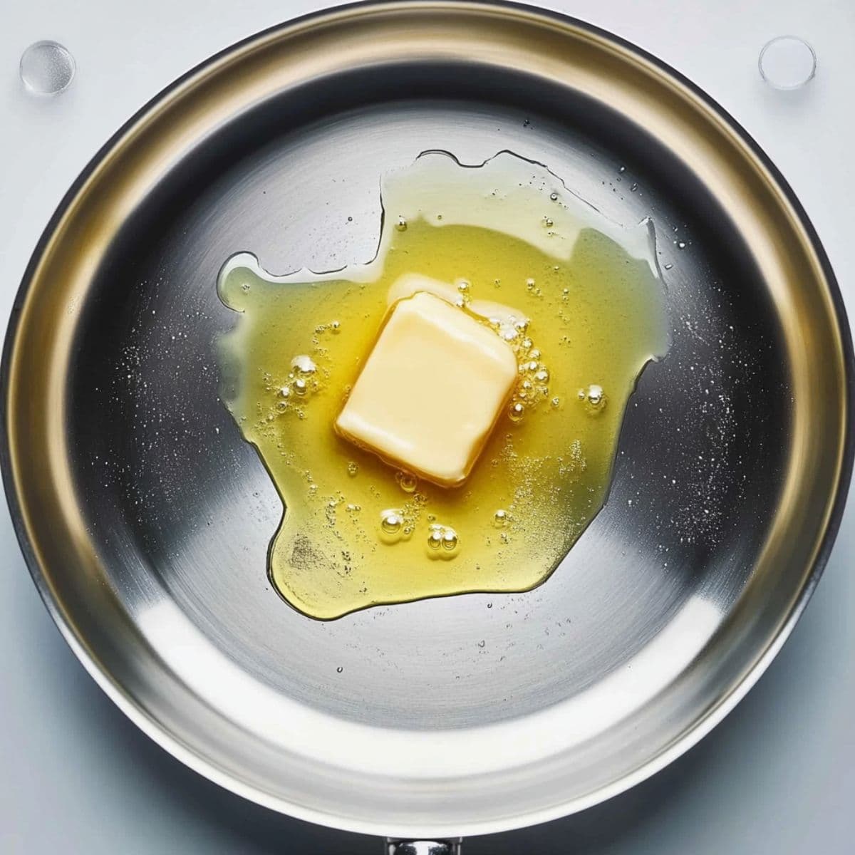 Top-down view of a cube of butter melting in a stainless steel pan on a white background.