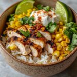 Street corn chicken rice bowl featuring golden seasoned chicken slices, bright yellow corn kernels, fluffy white rice, creamy dressing, fresh cilantro, and lime wedges in rustic ceramic bowl