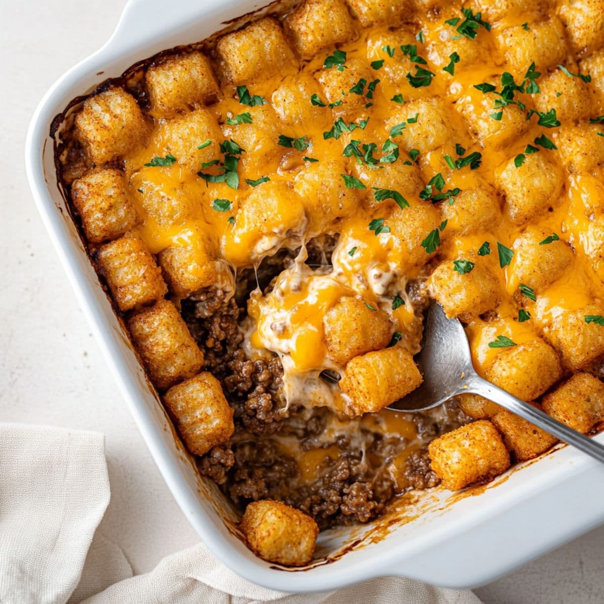 A top-down view of a tater tot casserole in a white baking dish, with crispy golden tater tots arranged neatly on top. A metal spoon is lifting a portion to reveal melted cheddar cheese, creamy sauce, and seasoned ground beef underneath, garnished with chopped parsley.