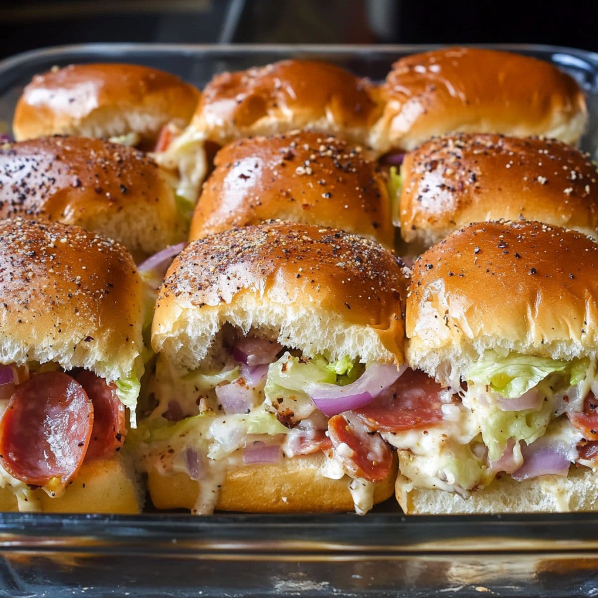 Tray of fresh Italian sliders with melted cheese, pepperoni, shredded lettuce, and red onions, served warm inside golden-baked Hawaiian rolls.