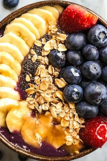 Acai bowl recipe with vibrant purple base topped with banana slices blueberries strawberries and granola