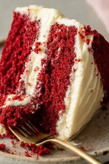 Classic red velvet cake slice with fluffy red layers and cream cheese frosting on ceramic plate with gold fork