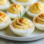 Classic deviled eggs garnished with fresh dill and paprika on white plate, showing creamy yellow filling and perfect presentation