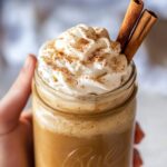 Creamy pumpkin spice latte in mason jar with whipped cream and cinnamon sticks, held in hands showing rich orange color and fall spices