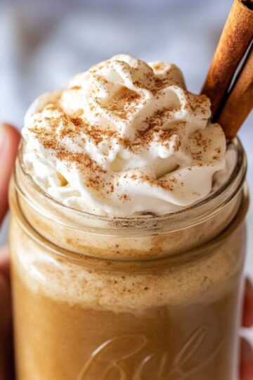 Creamy pumpkin spice latte in mason jar with whipped cream and cinnamon sticks, held in hands showing rich orange color and fall spices