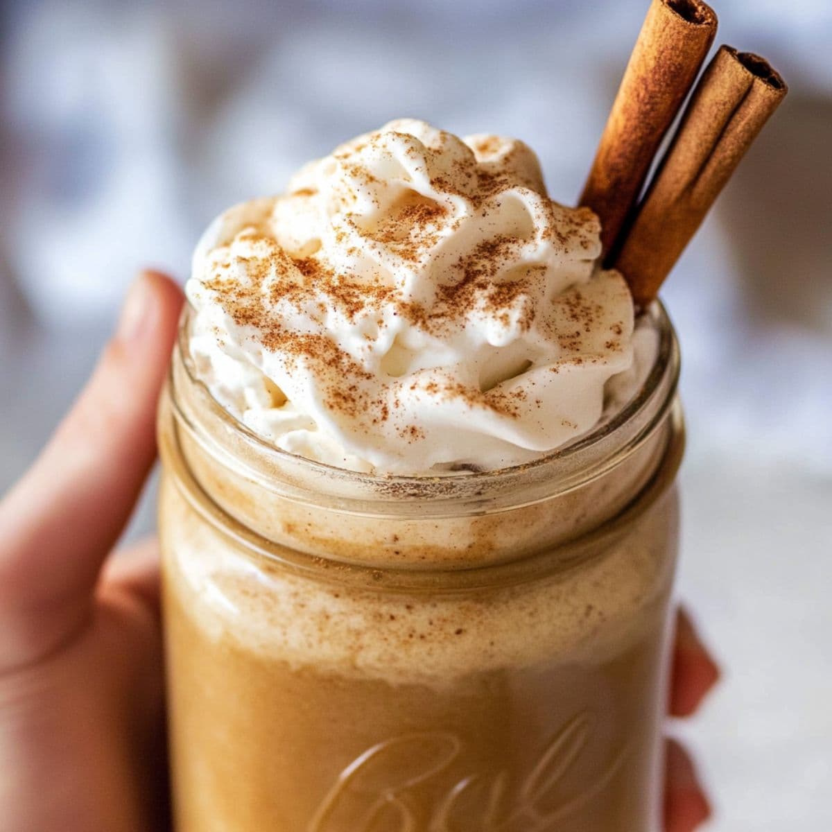Creamy pumpkin spice latte in mason jar with whipped cream and cinnamon sticks, held in hands showing rich orange color and fall spices