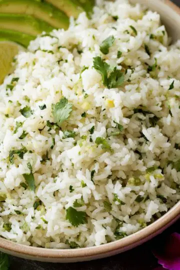 Cilantro lime rice served in a wooden bowl with fresh lime wedges, sliced avocado, and cilantro garnish on a dark surface
