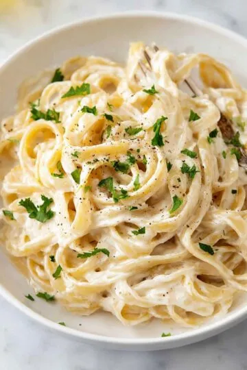 Alfredo sauce recipe showing creamy fettuccine pasta in white bowl with fresh parsley, black pepper and fork twirling noodles coated in silky homemade sauce