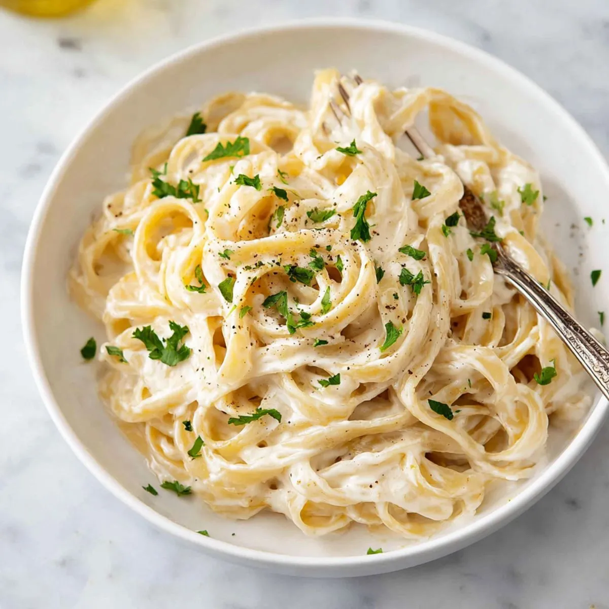 Alfredo sauce recipe showing creamy fettuccine pasta in white bowl with fresh parsley, black pepper and fork twirling noodles coated in silky homemade sauce