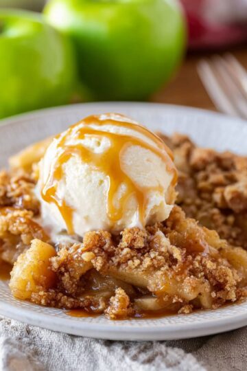 Apple crumble served on white plate with vanilla ice cream and caramel drizzle showing golden buttery topping and tender cinnamon apples with fresh green apples in background