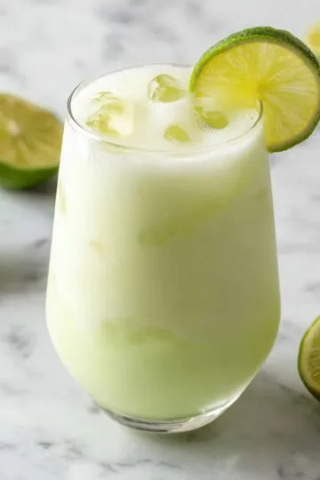 Brazilian lemonade in a tall glass with creamy pale green color garnished with fresh lime wheel and ice cubes with halved limes scattered on white marble background