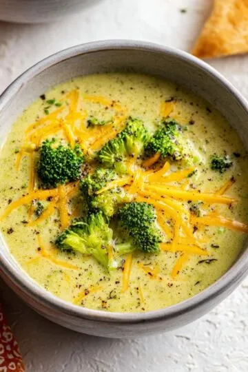 Broccoli cheddar soup in a ceramic bowl with melted cheddar cheese and bright green broccoli florets floating in creamy golden soup base with black pepper sprinkled on top and crusty bread in background