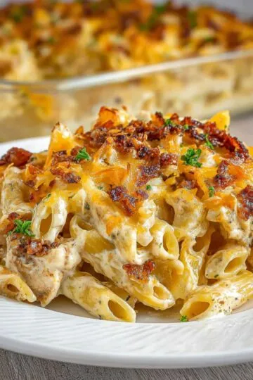 Chicken bacon ranch pasta recipe on white plate showing creamy rigatoni noodles with crispy bacon bits, melted cheese and fresh herbs with casserole dish in background for ultimate comfort food dinner