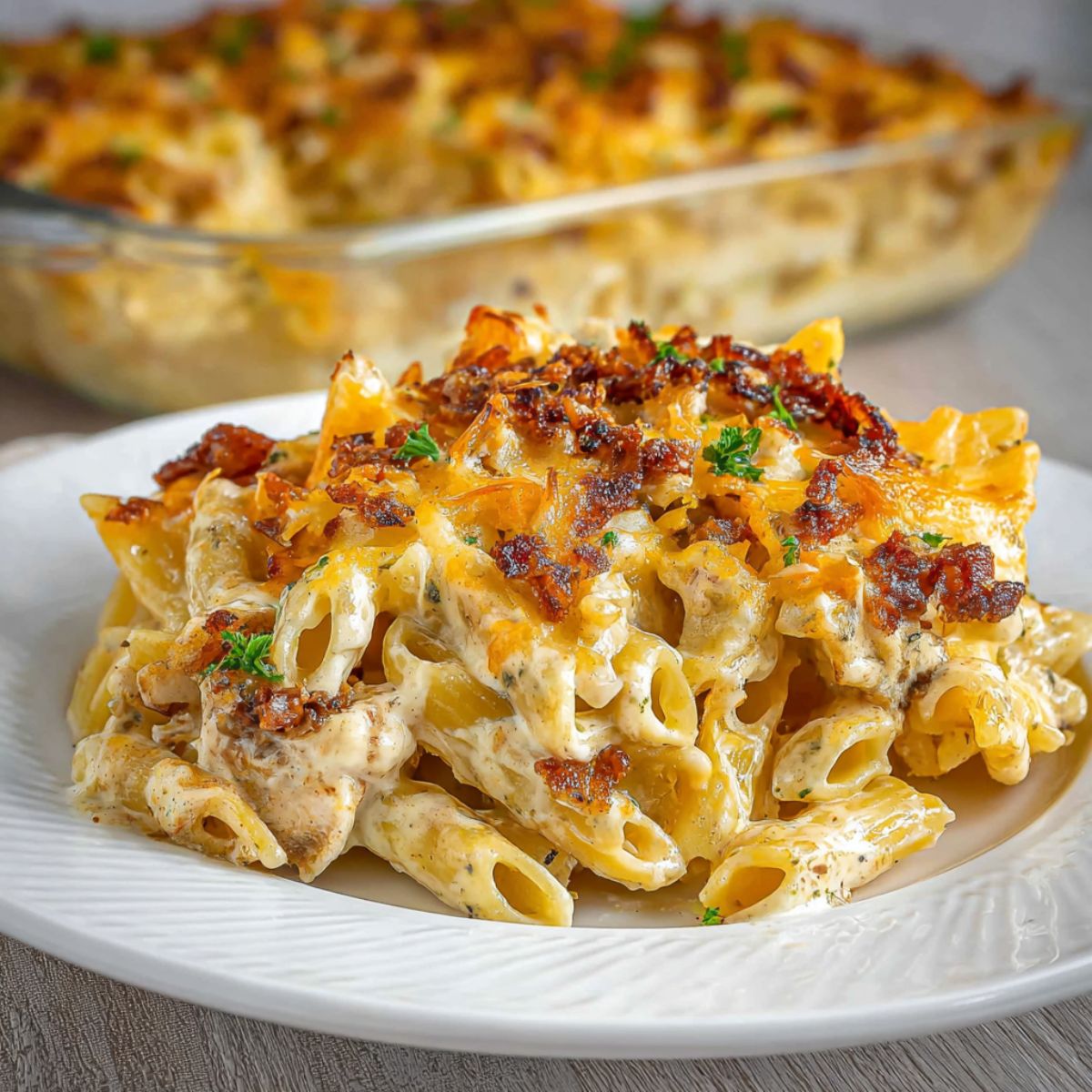 Chicken bacon ranch pasta recipe on white plate showing creamy rigatoni noodles with crispy bacon bits, melted cheese and fresh herbs with casserole dish in background for ultimate comfort food dinner