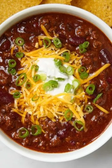 Chili recipe in white bowl topped with shredded cheese, sour cream, and green onions with tortilla chips on the side