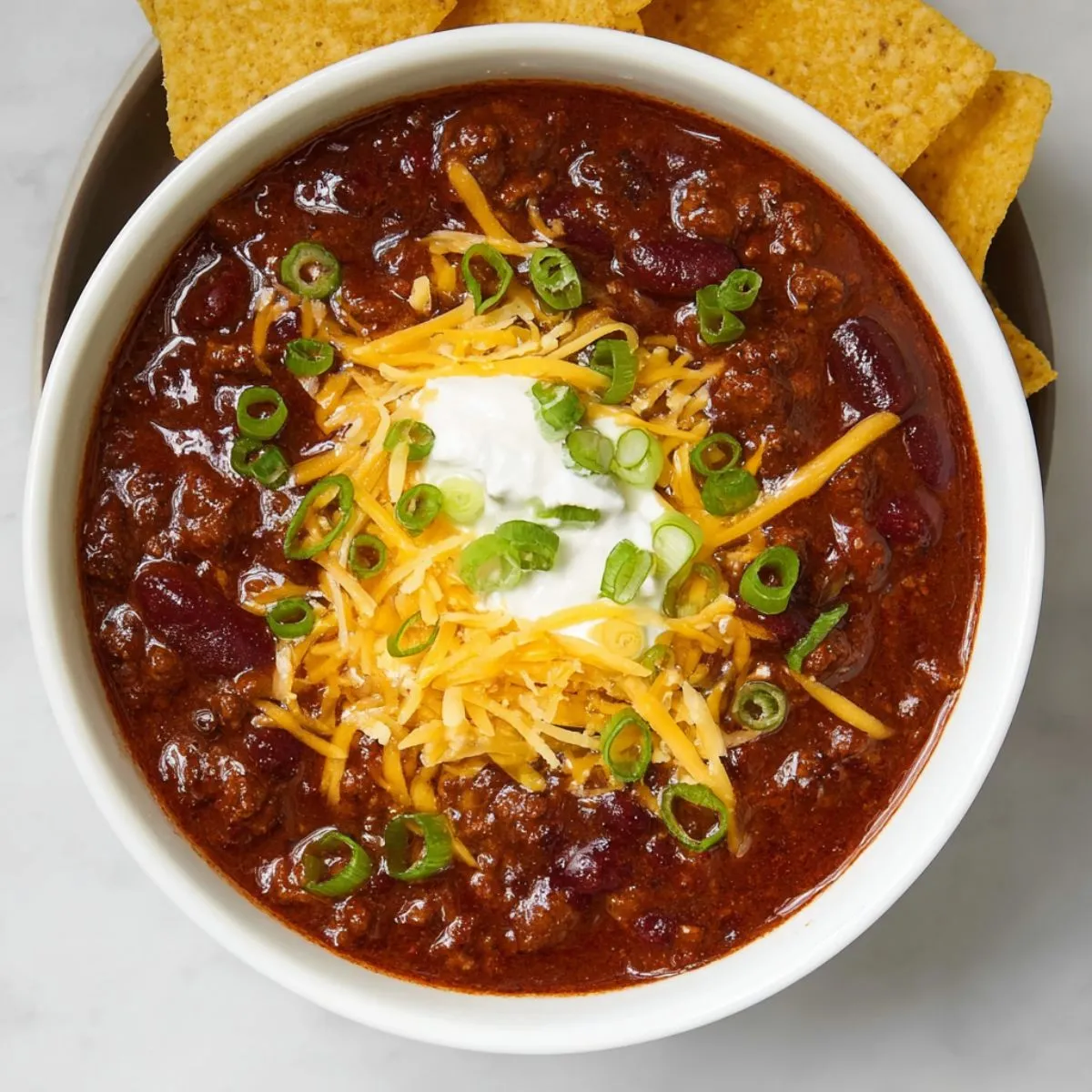 Chili recipe in white bowl topped with shredded cheese, sour cream, and green onions with tortilla chips on the side