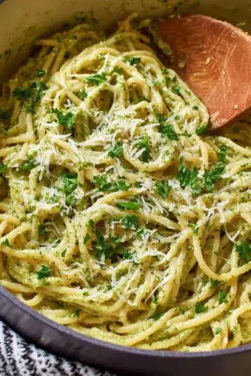 Pesto pasta in dark skillet with wooden spoon showing linguine noodles coated in vibrant green basil pesto sauce with fresh parsley and grated Parmesan cheese