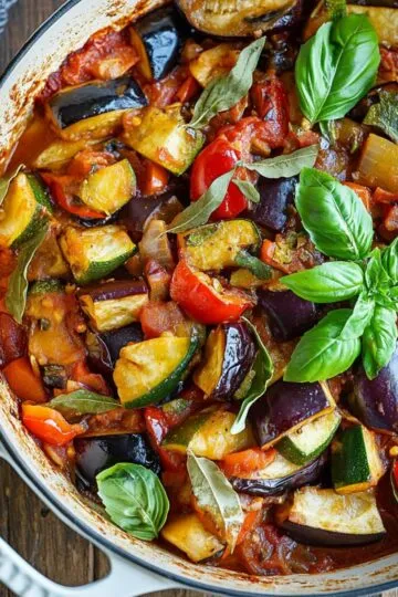 Ratatouille recipe in white cast iron pan with colorful eggplant, zucchini, red peppers, tomatoes and fresh basil leaves
