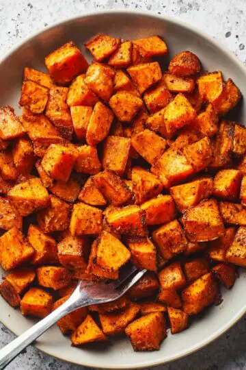 Roasted sweet potatoes cubes with caramelized edges and golden brown color seasoned with herbs in white ceramic serving bowl with silver fork
