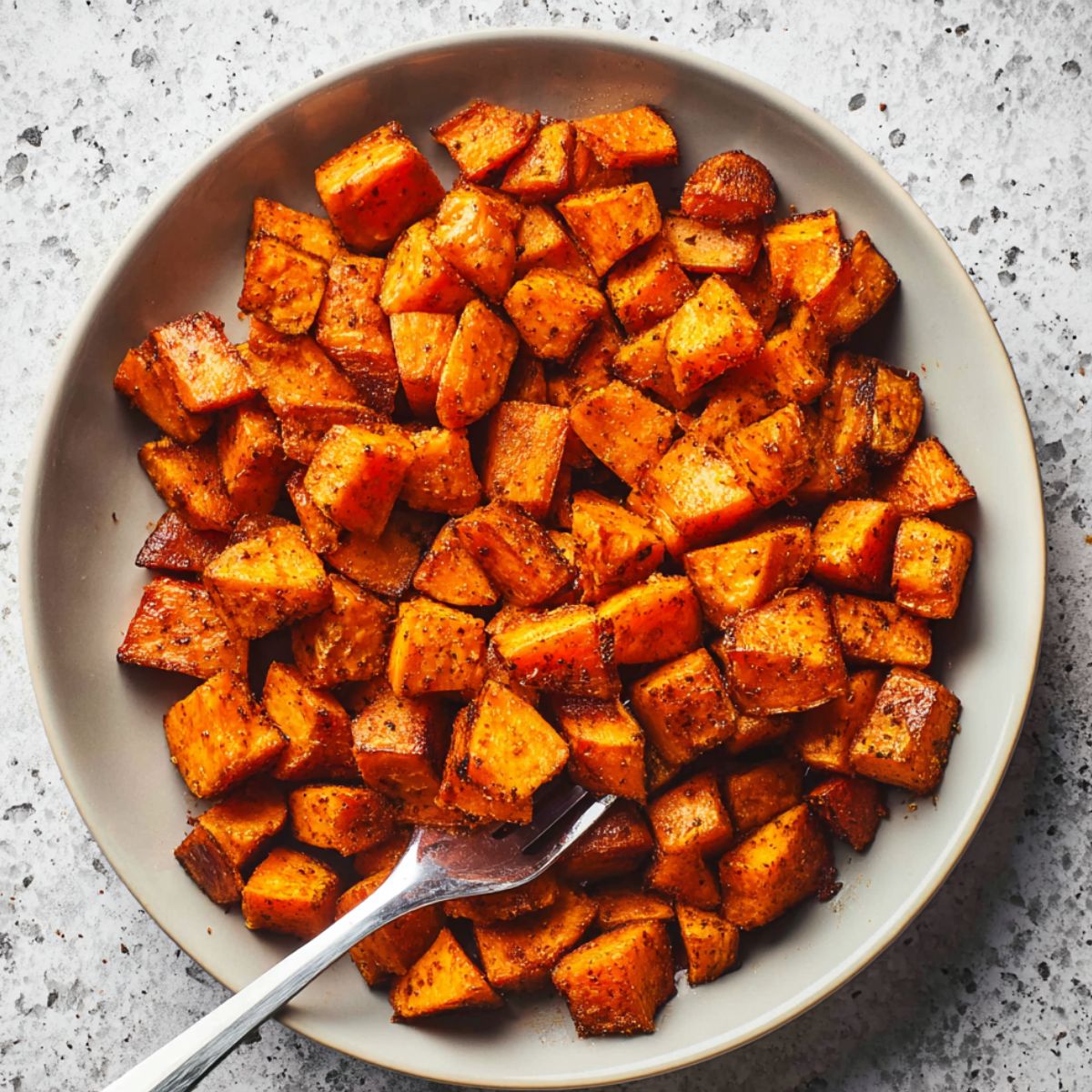 Roasted sweet potatoes cubes with caramelized edges and golden brown color seasoned with herbs in white ceramic serving bowl with silver fork