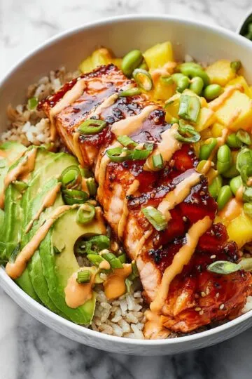 Salmon bowl with glazed teriyaki salmon fillet over brown rice topped with avocado slices edamame pineapple and spicy mayo sauce in white ceramic bowl